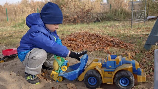 Young child wearing a blue jacket and hat engrossed in playing with a toy cement mixer and a large front loader truck in a sandy playground during a cool autumn day outdoors