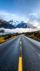 A road leads towards majestic snow-capped mountains, partially obscured by mist, under a cloudy blue sky