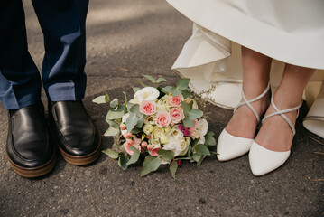 On their wedding day, the bride and groom stand on an asphalt road with a bouquet of flowers between their feet