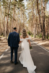 On their wedding day, the bride and groom hold hands and walk along an asphalt road in a pine forest