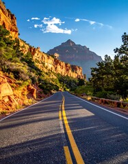 A road winds through a canyon, flanked by red rock cliffs and trees under a blue sky