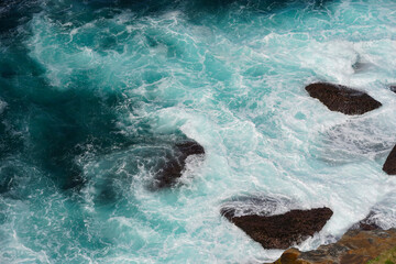Waves crashing against a cluster of dark brown rocks. The water is a stunning mix of deep blue and...