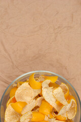 An overhead shot captures a clear glass bowl filled with small, rough pieces of yellow mandarin peel with white pith attached, resting on a background of wrinkled brown craft paper with ample copy spa