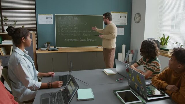 Medium full shot of cheerful middle-aged male Caucasian teacher standing by blackboard with AI prompting algorithm, holding tablet, explaining new topic to class of diverse students with laptops