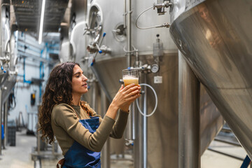 Woman brewer evaluating beer sample, producing craft beer in a modern brewery with fermentation tanks