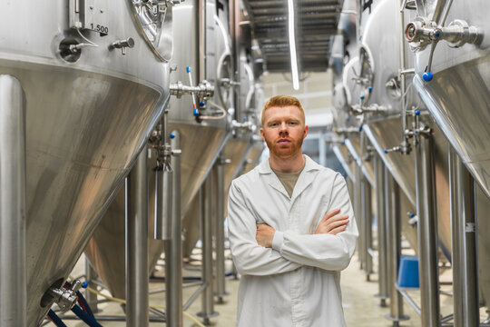 Man in lab coat with arms crossed stands confidently before stainless steel fermentation tanks in a modern craft brewery production plant - Powered by Adobe