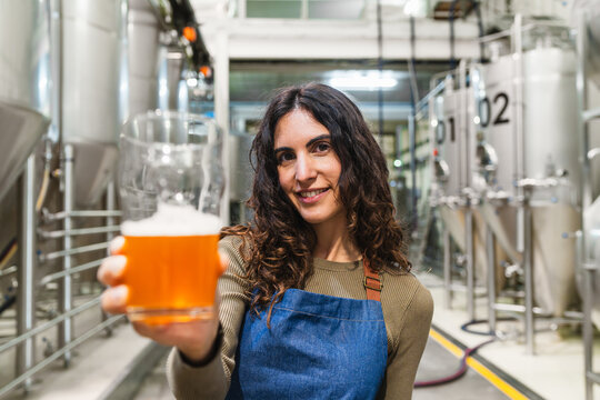 Woman holding a glass of craft beer, smiling, working in a modern brewery with large stainless steel tanks