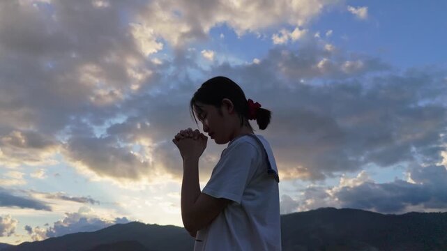 A girl praying to God with holding hands and closing her eyes at sunset shining brightly sky background.	