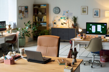 Modern open plan office workspace showing empty desks with laptops, monitors, office supplies and plants, background featuring organized shelves and digital screens in contemporary setting