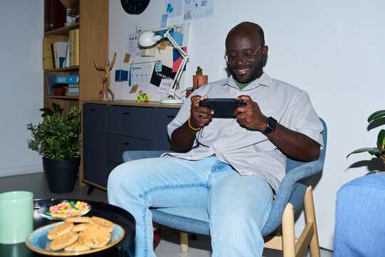 Black man sitting in modern office chair playing handheld gaming console smiling, surrounded by plants and snacks on table, business charts visible on wall