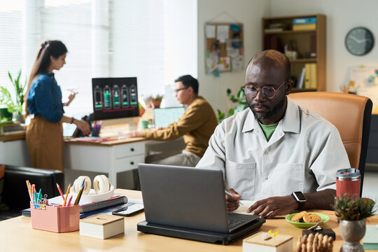 Black man working on laptop at desk in modern office, multiethnic colleagues collaborating in background, digital devices and office supplies visible on workspaces - Powered by Adobe