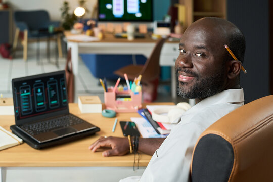 Portrait of Black man sitting at desk smiling at camera, holding pencil behind ear, working on laptop with smartphone interface designs displayed on screen in modern office - Powered by Adobe