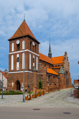 Obraz premium A historic red-brick church with a clock tower and a traditional tiled roof in Tolkmicko, Poland. The photo, taken on a sunny summer day, highlights the building's architecture.