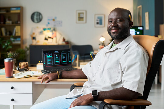 Portrait of Black man sitting at desk smiling at camera, working on laptop with smartphone interface designs on screen, wearing smartwatch in modern office