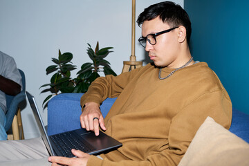 Asian man sitting on sofa using laptop with focused expression, wearing glasses and chain necklace,...