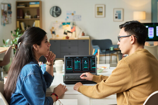 Asian man showing smartphone interface designs on laptop to woman during collaborative discussion in modern office workspace