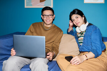 Hispanic man using laptop while sitting on couch next to Hispanic woman holding smartphone and wearing headphones, both looking toward camera and smiling