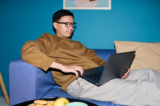 Man reclining on sofa using laptop, wearing glasses and focusing on screen, sitting in relaxed posture with snacks and fruit on table in foreground