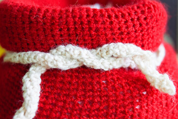 A bright close-up of a red knitted Christmas gift bag tied with a white knitted ribbon. The detailed texture of the yarn and festive colors create a cozy, holiday-themed composition perfect for season