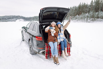 Two young women take selfies with phones while sitting in trunk of car on sunny winter day. Beautiful friends have fun, blog, enjoy road trip through snowy forest. Concept of friendship, adventure.