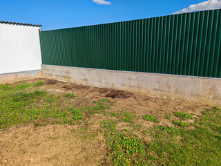 Green corrugated tin fence in the garden. Old concrete wall before paining