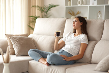 Fototapeta premium Pensive relaxed woman sitting on a couch holding coffee cup