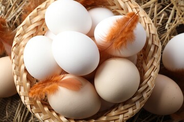 Raw chicken eggs in wicker basket and feathers on straw, top view