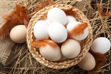 Raw chicken eggs in wicker basket, feathers and straw on table, flat lay
