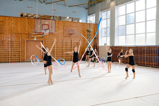 Group of Caucasian teenage girls performing rhythmic gymnastics routine with hoops and ribbons in sports hall, each gymnast executing synchronized movements on gym floor - Powered by Adobe