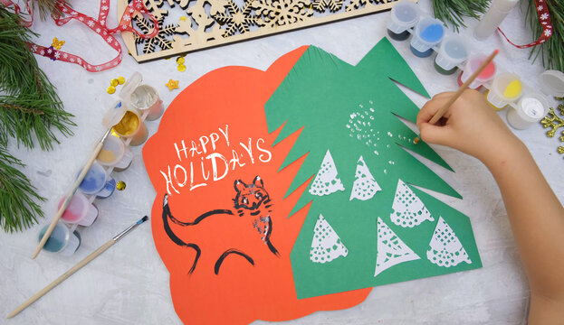 Child making a greeting card from colorful paper, paintbrush. Happy holidays text. Craft for children. Nice greeting card showing  Christmas snowy forest. Elementary. Top view - Powered by Adobe