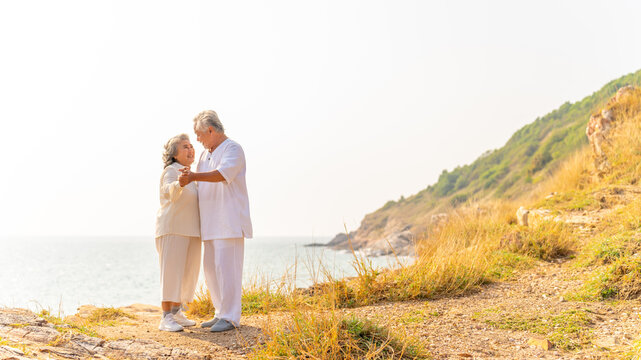 Happy Asian family senior couple dancing together on coastline at sunset. Healthy elderly husband and wife enjoy outdoor lifestyle travel nature at the sea in summer. Old people mental health wellness - Powered by Adobe