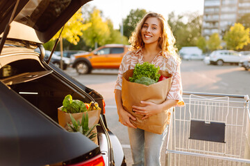 Smiling woman stands in parking lot with bag of groceries next to car in sunny parking lot. Beautiful woman with cart full of fresh fruits, vegetables loads groceries into car. Concepts of shopping.