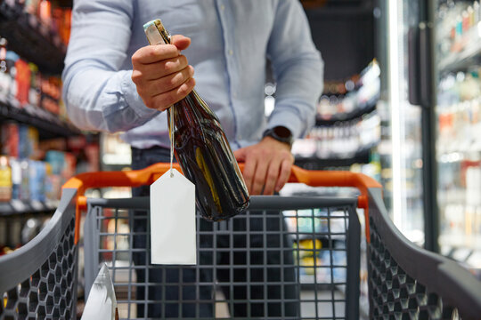A man is pushing a shopping cart that contains a bottle of wine inside it