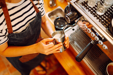 An experienced barista prepares freshly brewed coffee in a cozy cafe. A young woman froths and pours milk into an espresso. Small business concept, beverages.