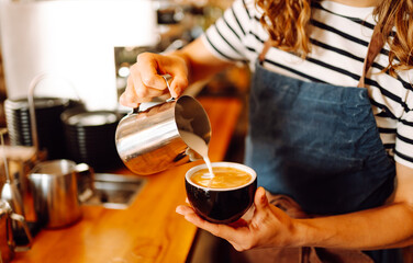 An experienced barista prepares freshly brewed coffee in a cozy cafe. A young woman froths and...