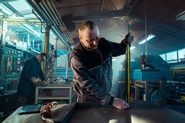 Metalworker measuring metal sheet in workshop with colleague welding