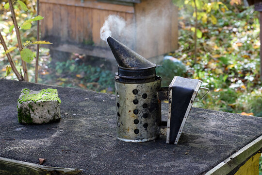 A beekeeping smoker emits smoke while placed on a textured wooden surface next to a mossy stone, complemented by a blurred outdoor environment with a beehive in the background.