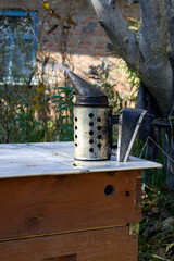 A close-up view of a classic bee smoker positioned on a wooden beehive in an outdoor setting, highlighting the tools of beekeeping and the peacefulness of rural life.