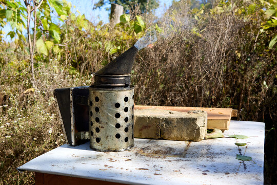 A bee smoker emitting smoke stands on a weathered table, accented by foliage and beekeeping tools, displaying the beekeeping practice with rustic, natural charm, and outdoor setting. - Powered by Adobe
