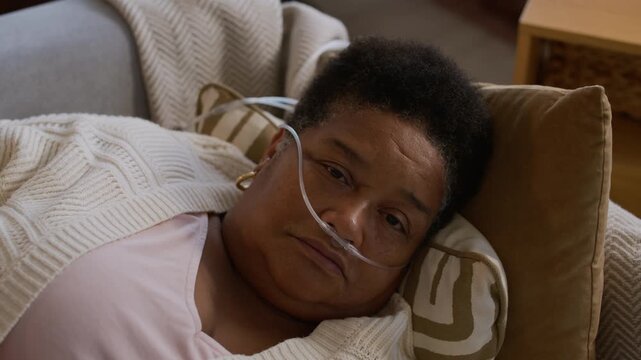 Medium close up portrait of elderly woman with nasal cannula getting home treatment lying on sofa during therapy and looking at camera