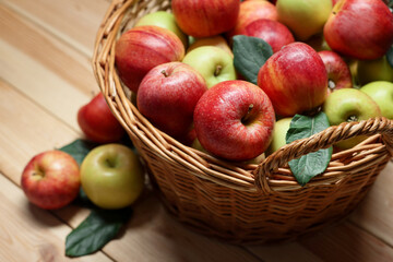 Fresh apples in wicker basket and green leaves on wooden table, closeup
