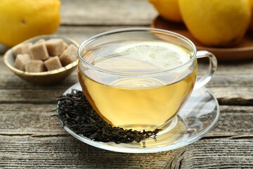 Aromatic black tea in glass cup, dried leaves, brown sugar and lemons on wooden table, closeup