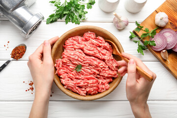 Woman stirring raw minced meat in bowl at white wooden table, top view