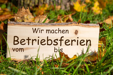 Betriebsferien Herbst: Holzschild mit hinzugefügtem Text WIR MACHEN BETRIEBSFERIEN VOM/BIS auf einer Wiese mit Herbstlaub