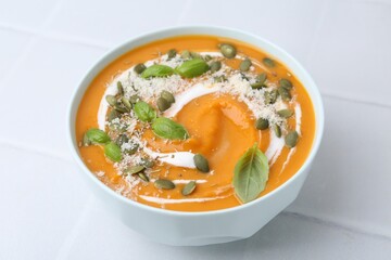 Delicious pumpkin soup with spices, cheese and sour cream in bowl on white tiled table, closeup