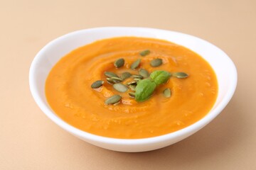 Delicious pumpkin soup with seeds and basil in bowl on beige table, closeup
