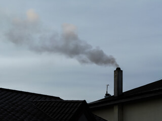 Smoke rising from a chimney on a residential roof against a cloudy sky, symbolizing heating, pollution, or winter atmosphere