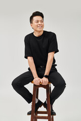 Handsome young Asian man sitting on a wooden stool with a joyful expression in a studio setting