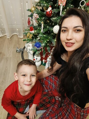 A woman and a boy sitting on the floor in front of a decorated Christmas tree. Garlands, ornaments, and baubles glitter on the tree. The comfort and joy of a family holiday