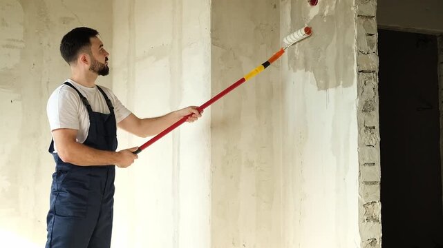 A bearded man in a work uniform is priming the walls after applying gypsum plaster